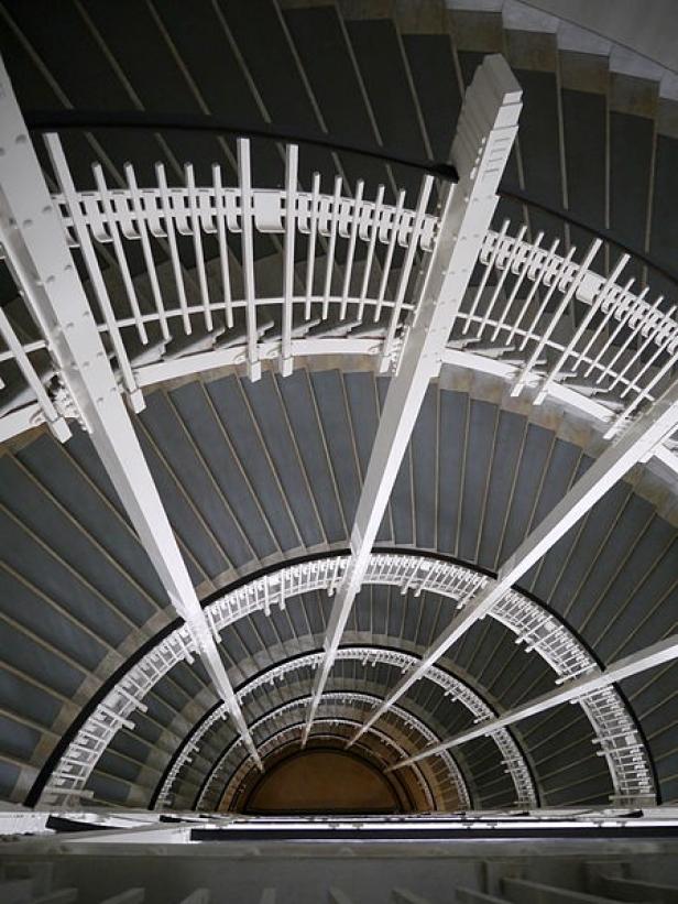 Blick von oben auf ein spiralförmiges Treppenhaus mit grau gestrichenen Stufen und weißem Geländer.