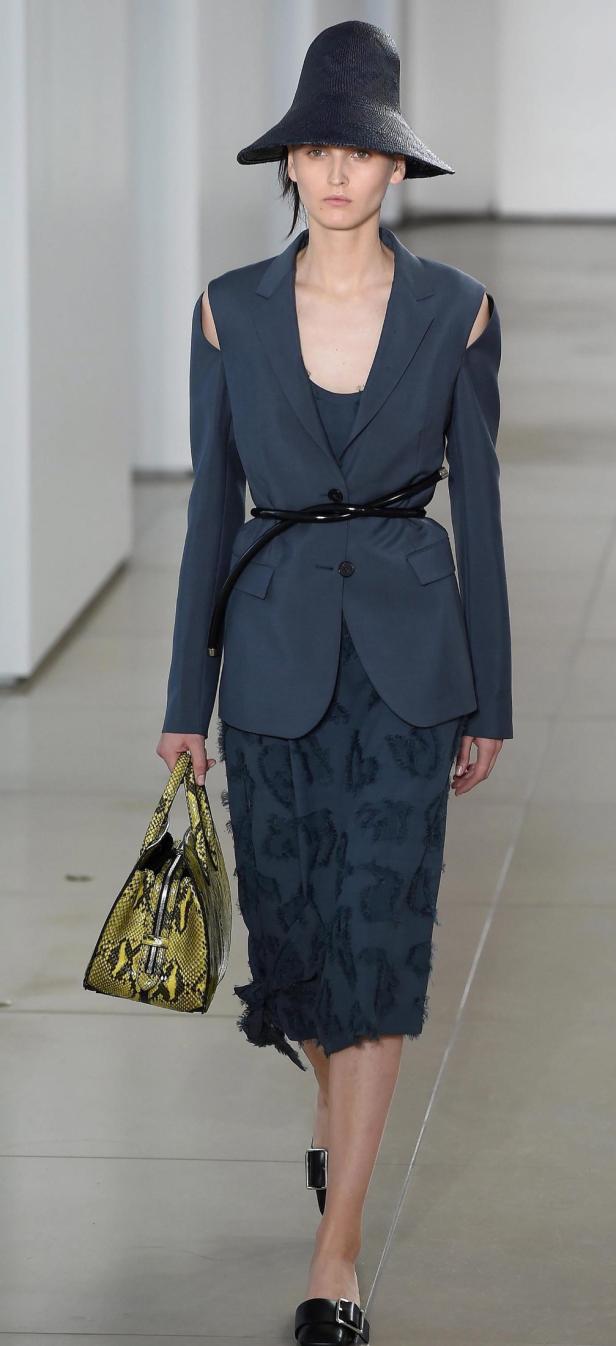 Eine Frau mit Hut und blauem Blazer mit Cut-Outs auf dem Laufsteg präsentiert eine Handtasche mit Schlangenmuster.