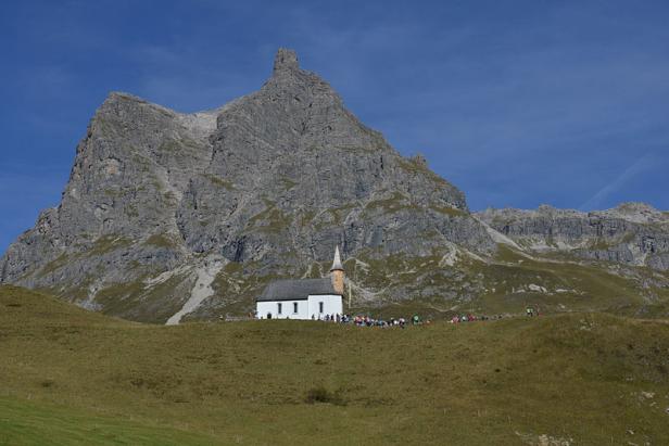 Eine kleine Kirche vor einer markanten Bergkulisse mit Menschen im Vordergrund.