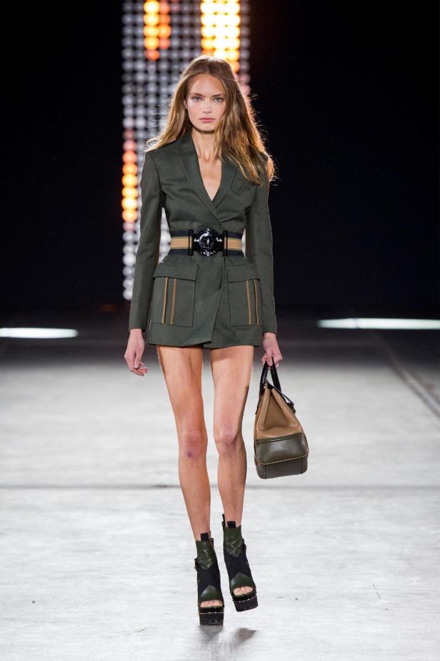 Eine Frau auf dem Laufsteg trägt ein kurzes, olivgrünes Blazerkleid mit Gürtel und eine Handtasche.