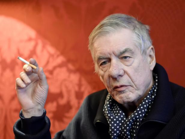 Ein älterer Mann mit einem gepunkteten Schal hält eine Zigarette in der Hand.