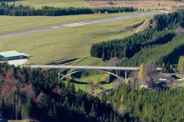 Eine Brücke überspannt ein Tal in einer bewaldeten Landschaft mit einer Landebahn im Hintergrund.