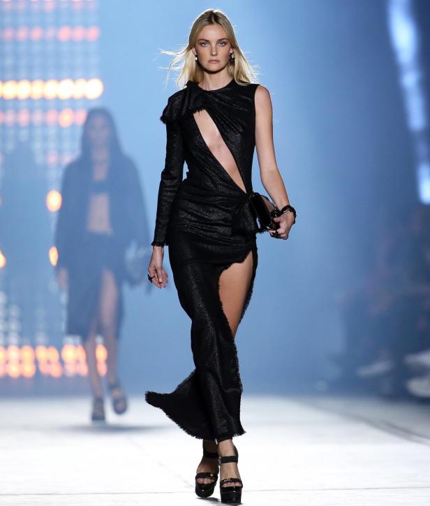 Eine blonde Frau in einem schwarzen, asymmetrischen Kleid auf einem Laufsteg.