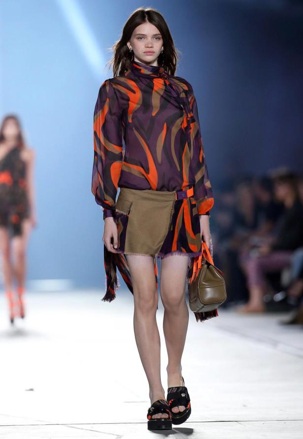 Ein Model auf dem Laufsteg trägt ein gemustertes Kleid und eine braune Handtasche.