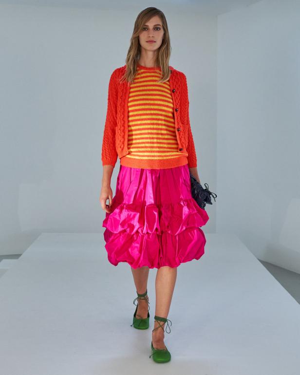 Eine Frau in einem orangefarbenen Cardigan, einem gestreiften Oberteil und einem pinkfarbenen Rock auf einem Laufsteg.