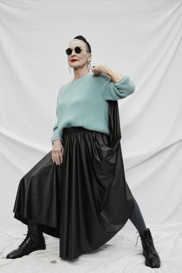 Eine ältere Frau mit Sonnenbrille posiert in einem türkisfarbenen Pullover und einem schwarzen Rock vor einem weißen Hintergrund.