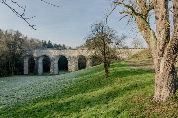 Eine Steinbogenbrücke überspannt eine grüne Wiese an einem sonnigen Tag.
