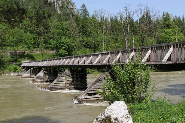 Eine hölzerne Brücke überspannt einen Fluss vor einer bewaldeten Landschaft.