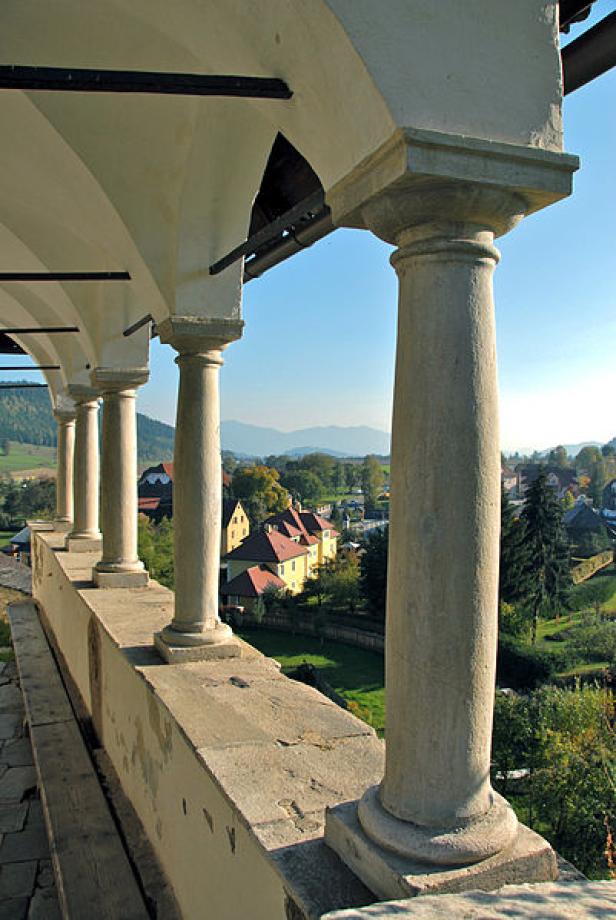 Blick durch die Säulen eines Bogengangs auf eine grüne Landschaft mit Häusern und Bergen im Hintergrund.