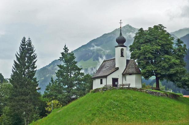 Eine kleine weiße Kapelle auf einem grünen Hügel vor einer Bergkulisse.