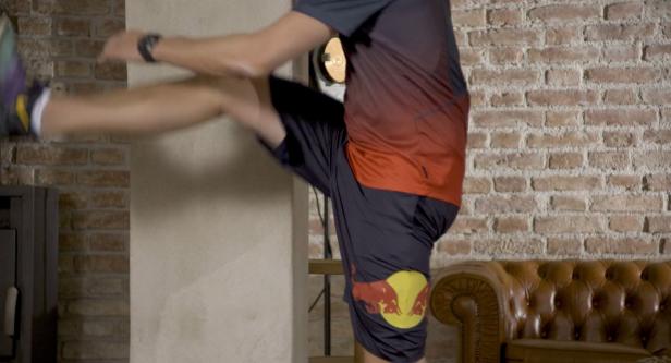 Ein Mann macht eine Dehnübung in einem Raum mit Backsteinmauer und Ledersofa.