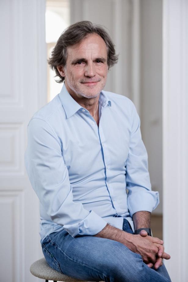 Ein Mann mit hellblauem Hemd und Jeans sitzt auf einem Hocker.