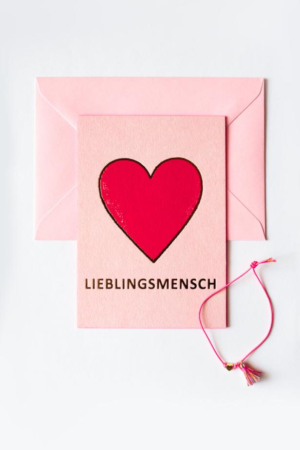 Eine rosa Karte mit einem roten Herz und der Aufschrift „Lieblingsmensch“ liegt neben einem Umschlag und einem Armband.