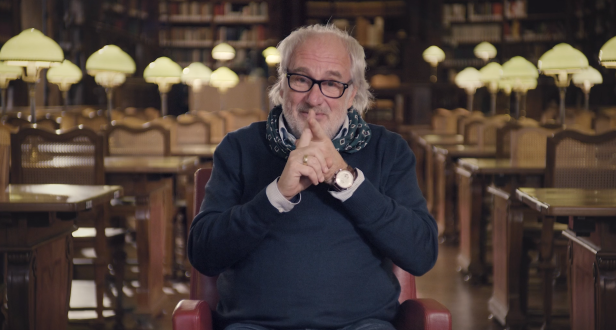 Ein Mann mit Brille sitzt in einer Bibliothek und hält die Hände vor dem Gesicht.