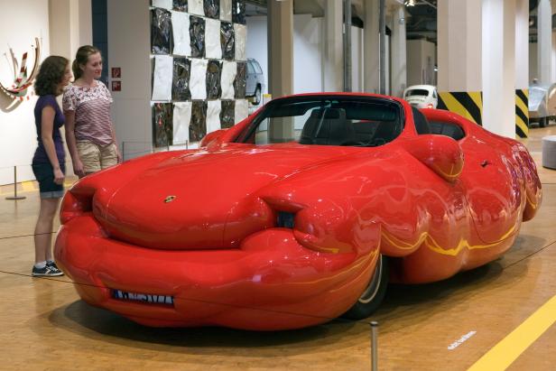 Zwei Frauen betrachten eine rote, überzeichnete Porsche-Skulptur in einer Ausstellung.