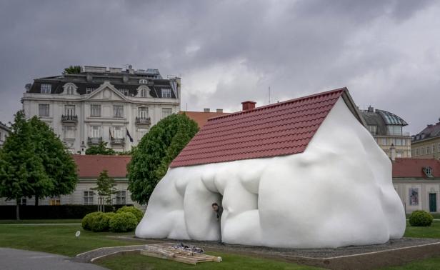 Ein ungewöhnliches, weißes Haus mit rotem Dach steht in einem Park, eine Person schaut aus dem Eingang.