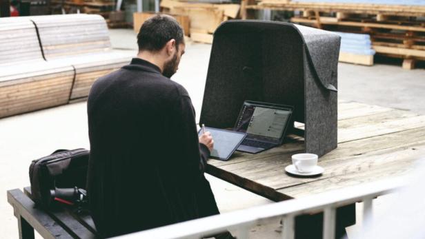 Ein Mann arbeitet an einem Laptop und einem Tablet in einer grauen Arbeitskabine im Freien.