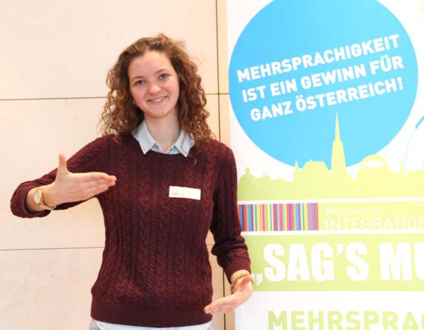 Eine junge Frau namens Anja steht vor einem Banner, das Mehrsprachigkeit bewirbt.