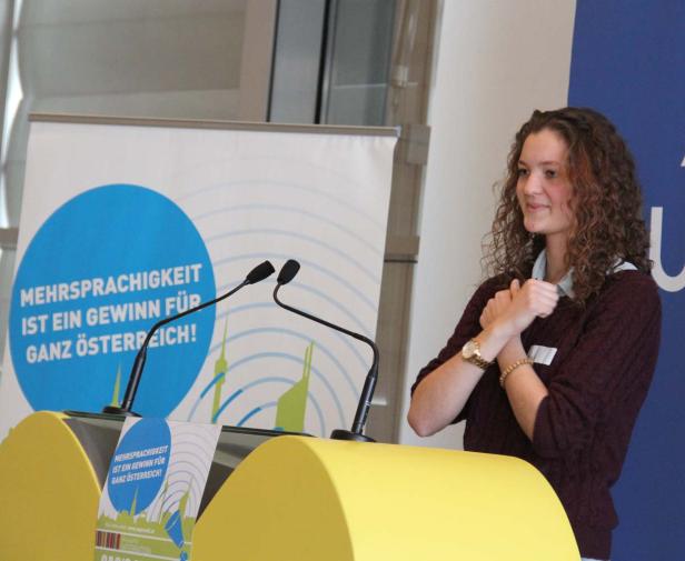 Eine junge Frau steht an einem Rednerpult mit der Aufschrift „Mehrsprachigkeit ist ein Gewinn für ganz Österreich!“.