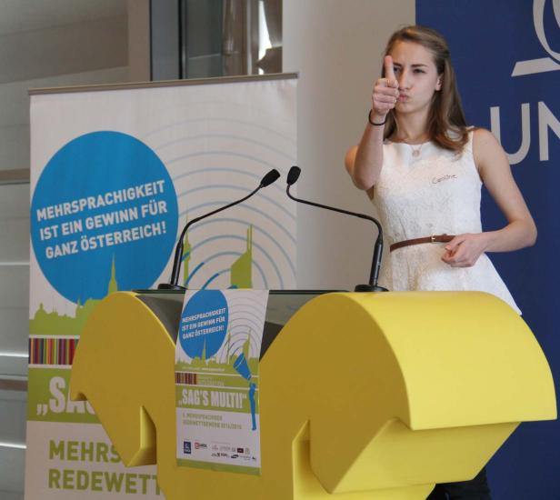 Caroline gibt bei einem Redewettbewerb vor einem gelben Rednerpult einen Daumen nach oben.