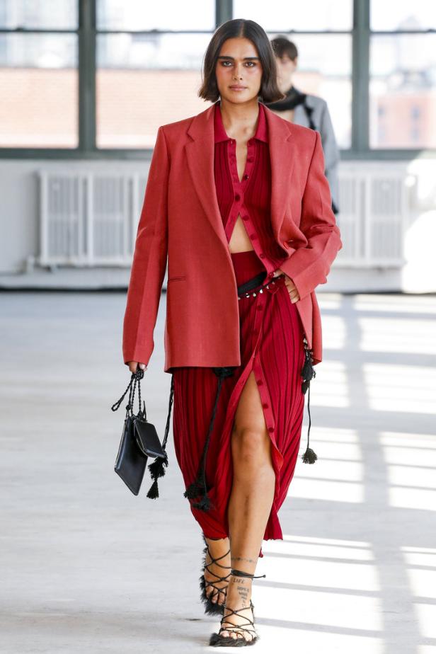 Eine Frau in einem roten Blazer und einem passenden Rock mit Schlitz läuft auf einem Laufsteg.