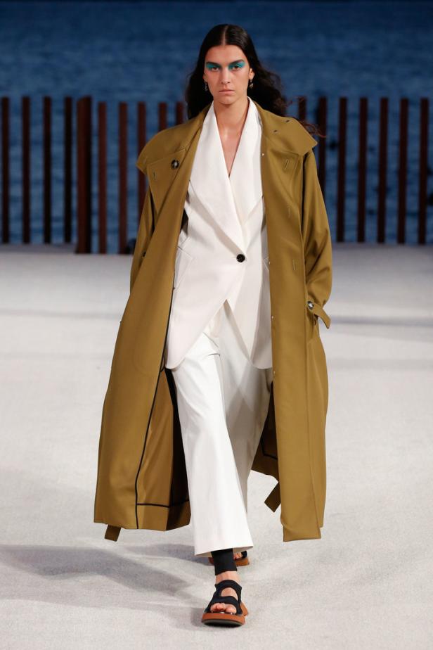 Eine Frau mit blauem Lidschatten trägt einen Trenchcoat über einem weißen Hosenanzug.