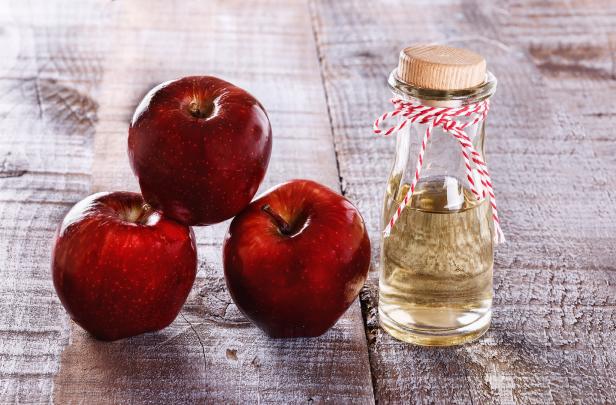 Drei rote Äpfel liegen neben einer Flasche Apfelessig auf einem Holztisch.