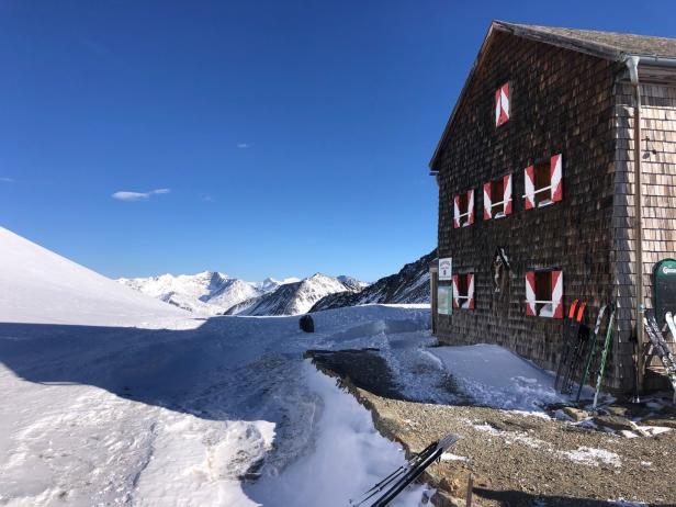 Eine Berghütte mit rot-weiß gestreiften Fensterläden vor einer verschneiten Berglandschaft.