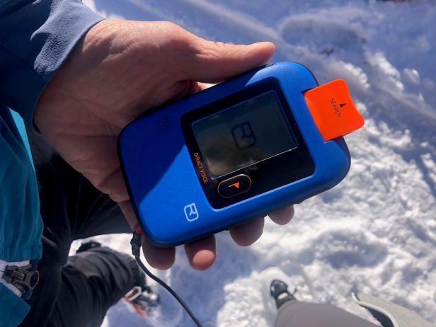 Eine Hand hält ein blaues Lawinenverschüttetensuchgerät (LVS) der Marke Ortovox im Schnee.
