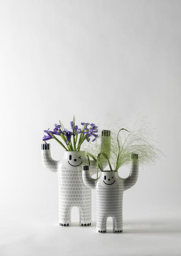 Zwei Vasen in Form von Figuren mit aufgemalten Gesichtern halten Blumen und Gräser.