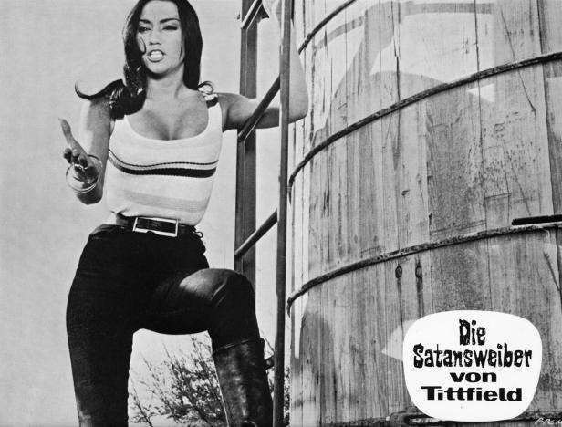 Eine Frau posiert vor einem Wasserturm mit dem Titel „Die Satansweiber von Tittfield“.