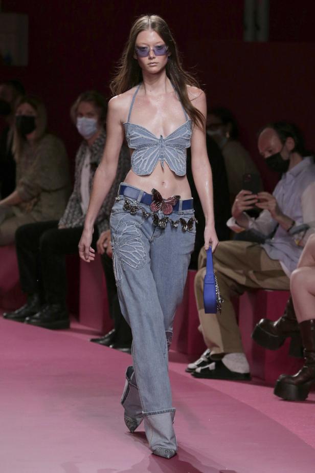Ein Model präsentiert ein Outfit mit Jeans, einem Oberteil in Schmetterlingsform und einer blauen Handtasche auf dem Laufsteg.