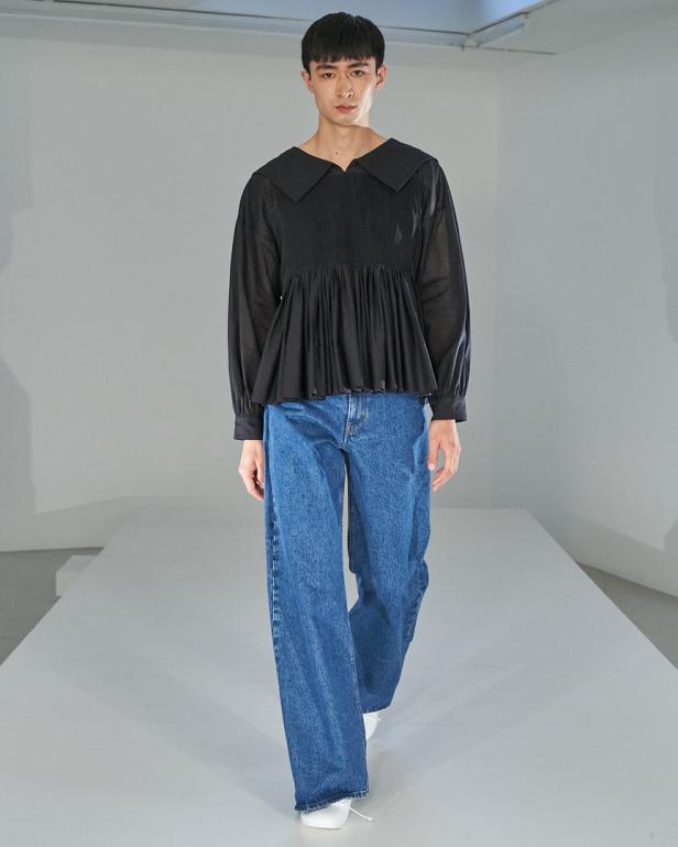 Ein schlanker Mann in einer schwarzen Bluse und blauen Jeans auf einem Laufsteg.