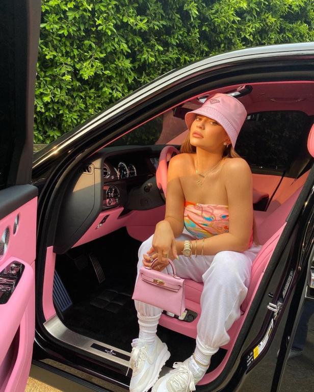 Eine Frau mit rosa Hut und Handtasche sitzt in einem Auto mit rosa Innenausstattung.