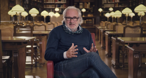Ein Mann mit Brille sitzt in einer Bibliothek und gestikuliert.