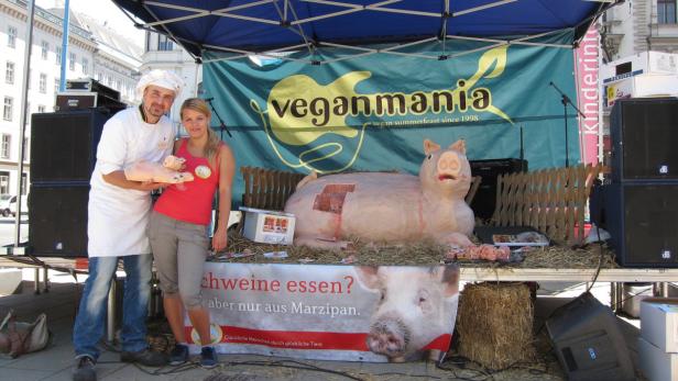 Ein Koch und eine Frau stehen vor einem Stand von „Veganmania“ mit einer großen Marzipanschwein-Skulptur.