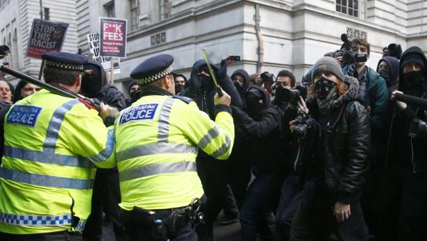 Polizisten stehen Demonstranten mit Schildern gegenüber, auf denen „Stop This Racist“ und „F**k Fees“ steht.