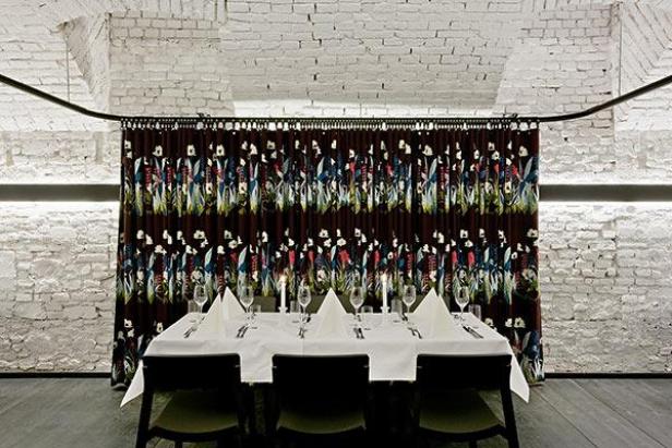 Ein gedeckter Tisch vor einer floral gemusterten Gardine in einem Raum mit Backsteinwänden.