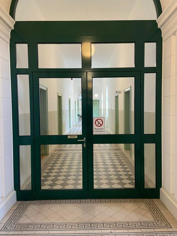 Eine grüne Doppeltür mit Glas führt in einen Flur mit Schachbrettmuster und einem "Rauchen verboten"-Schild.
