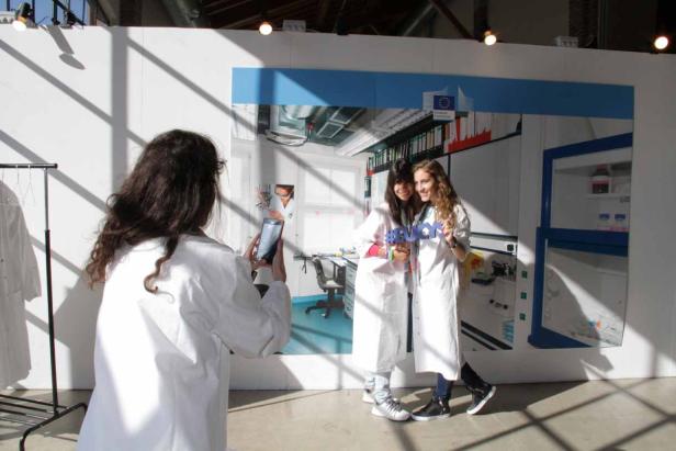 Zwei junge Frauen in Laborkitteln posieren vor einem Foto eines Labors.