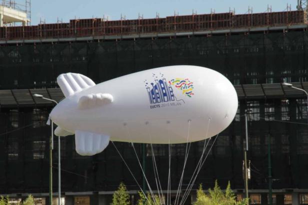 Ein weißer Werbe-Zeppelin mit dem Logo von EUCYS 2015 Mailand vor einem Gebäude.
