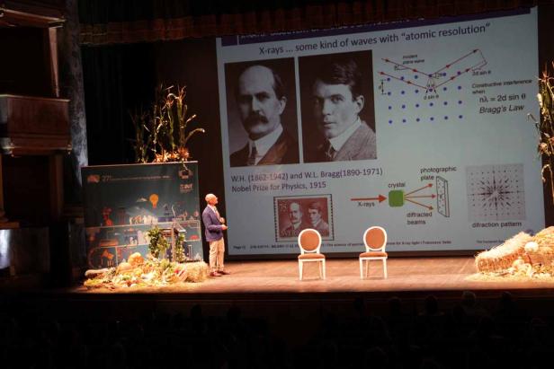 Ein Redner präsentiert auf einer Bühne Röntgenbeugung mit Porträts von W.H. und W.L. Bragg.