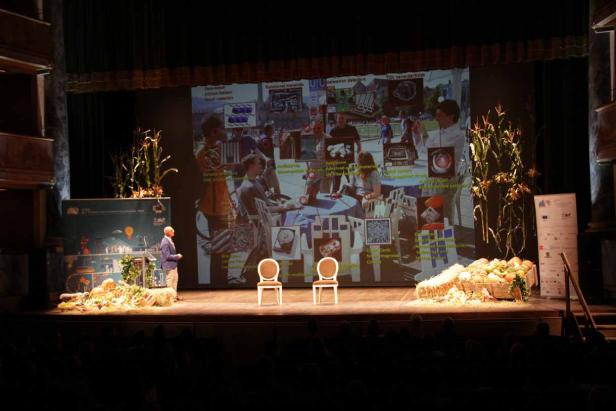 Ein Redner steht auf einer Bühne vor einer großen Leinwand mit einer Präsentation.