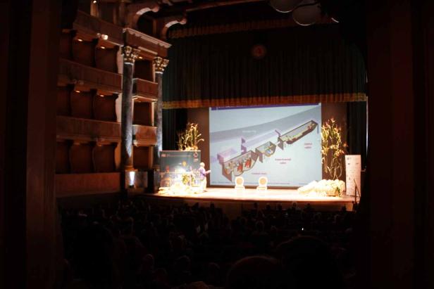 Eine Präsentation über eine wissenschaftliche Anlage auf einer Bühne vor Publikum.