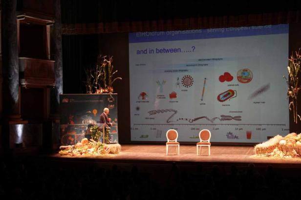 Ein Redner steht auf einer Bühne vor einer Präsentation über wissenschaftliche Organisationen.