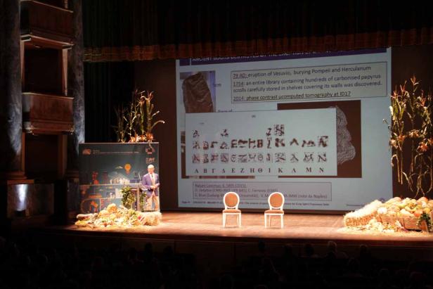 Ein Redner steht auf einer Bühne vor einer Präsentation über die Zerstörung von Herculaneum und Pompeji.