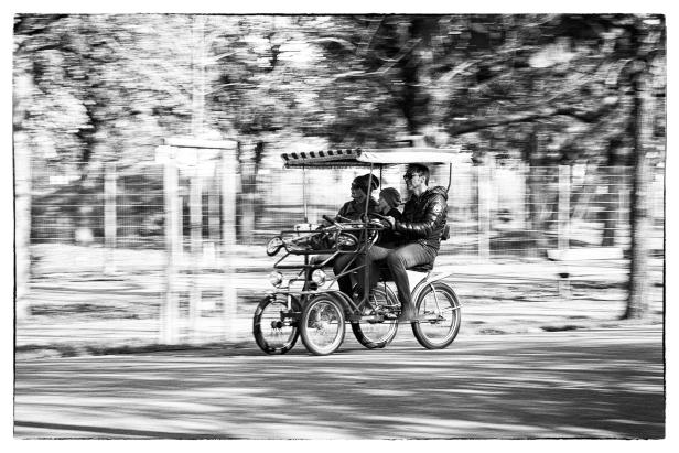 Drei Personen fahren mit einem überdachten Tandem-Fahrrad auf einer Straße.