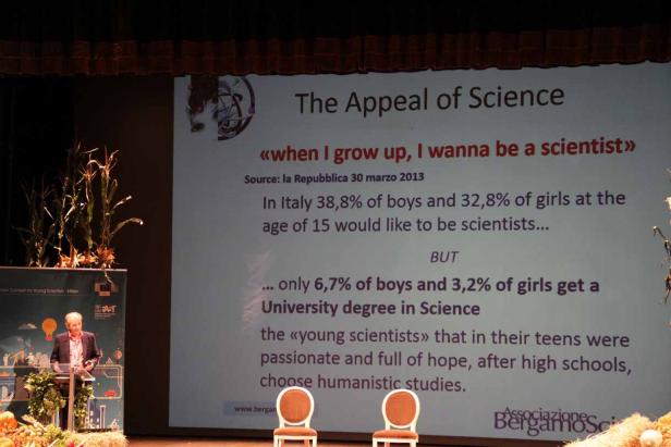 Ein Redner steht auf einer Bühne vor einer Präsentation über die Attraktivität der Wissenschaft.