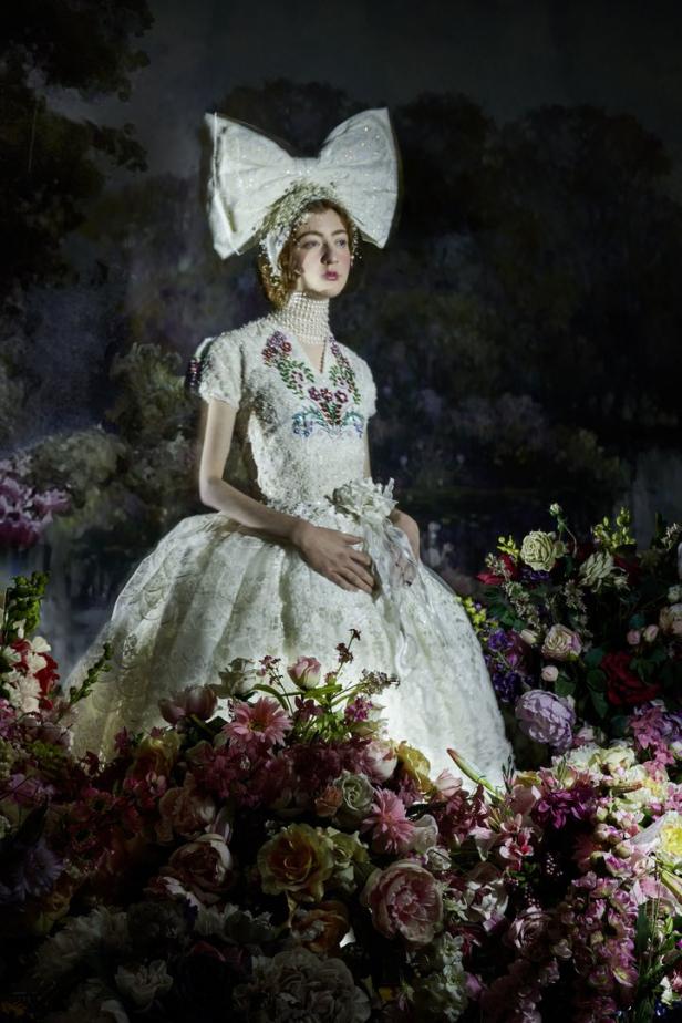 Eine Frau in einem weißen Kleid und einer großen Schleife steht inmitten von Blumen.
