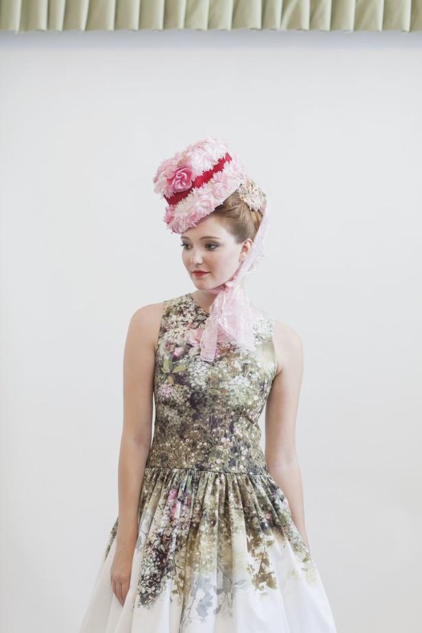 Eine Frau mit einem floralen Kleid und einem auffälligen Hut mit rosa Blumen.